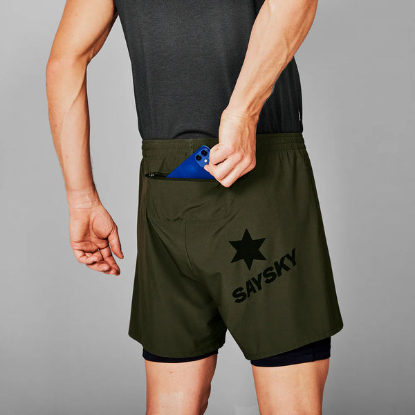 En person i mørk ærmeløs skjorte og Saysky 2 in 1 Pace Shorts 5'', Herre, Green, med SAYSKY- og stjernelogo, putter en blå telefon i shortsens baglomme med lynlås.