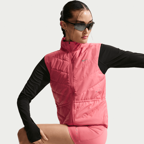 En kvinde iført Nike Swift Running Vest, Dame, i Sea Coral/Reflective Silver, sort langærmet top, pink shorts og solbriller poserer med en bøjet arm og tilbagestrøget hår på en almindelig baggrund.