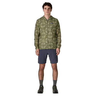 En person står og kigger fremad iført en Patagonia Houdini Jacket, Herre, Sand Waves/Caper Green, mørkegrå shorts, grå sokker og vandresko på en almindelig hvid baggrund.
