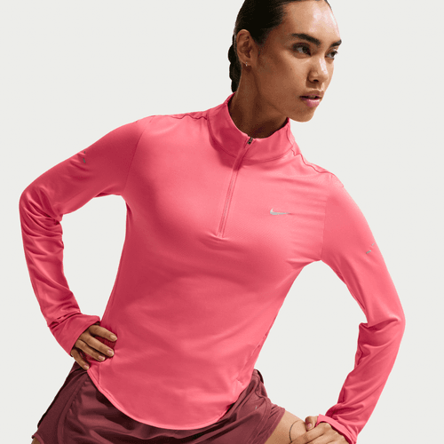 En kvinde i Nike Swift UV Half Zip Running Top, Dame, Sea Coral/Reflective Silv med reflekser og logo, kombinerer den med rødbrune shorts og poserer med hænderne i vejret og kigger til siden.