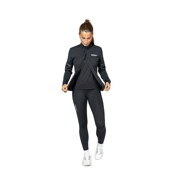 En kvinde i en sort Liiteguard Hydro-Tech løbejakke, sorte leggings og hvide sneakers står på en hvid baggrund, mens hun holder siderne af sin vandafvisende jakke og kigger ned.