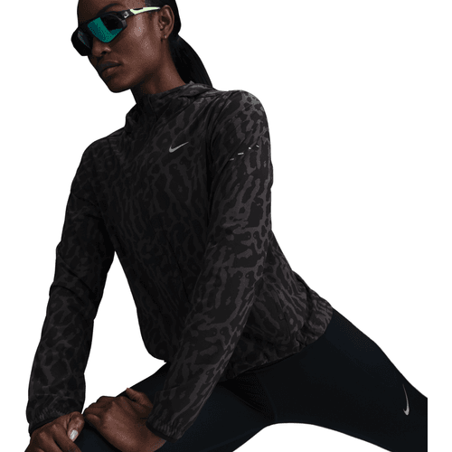 En kvinde strækker benet iført en sortmønstret Nike Dri-FIT løbejakke (Dame, Black/Anthracite/Reflective Silver) og sorte atletiske leggings. Hun bærer reflekterende solbriller og har håret trukket tilbage mod en hvid baggrund.