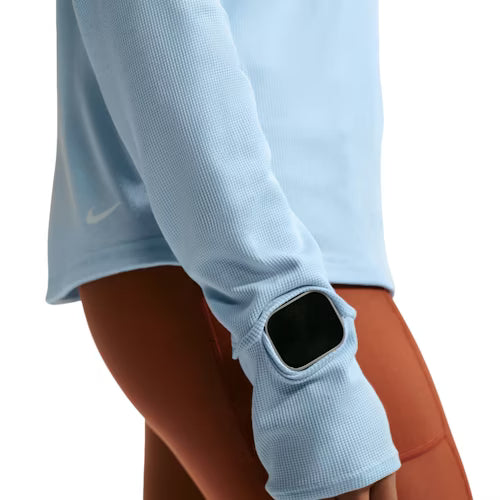 En person er iført en Nike ACG Half Zip Running Top i Hydrogen Blue/Summit White med huller til tommelfingrene og et urvindue sammen med højtaljede rustfarvede leggings. Kun overkroppen og armen er synlige.