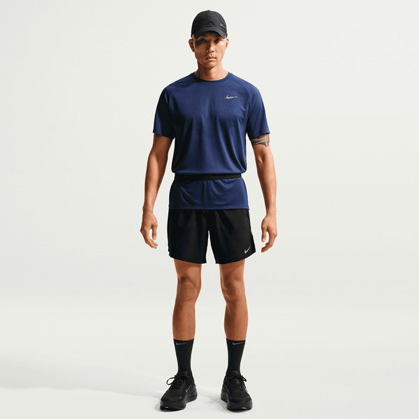 En mand ser frem for sig i en marineblå sportstrøje, Stride 7" Brief-Lined Running Shorts fra Nike i sort/reflekterende sølv, sorte sneakers, sorte sokker og en sort kasket. Baggrunden er helt hvid.
