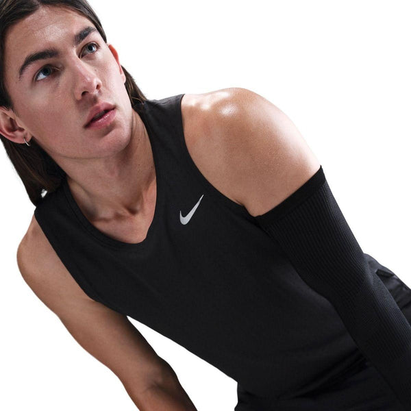 En person med langt brunt hår iført en ærmeløs sort Nike Dri-FIT Running Tank Top, Herre, Black/Reflective Silver og et sort kompressionsærme på højre arm kigger opad mod en almindelig hvid baggrund.