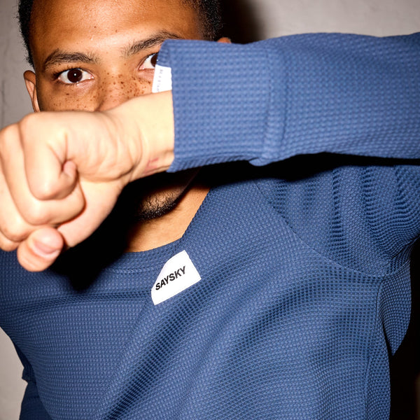 En person iført Saysky Motion Waffle Long Sleeve, Herre, Blue hæver armen og skjuler delvist ansigtet. Baggrunden er ensfarvet og lys-perfekt til løb eller aktive dage.