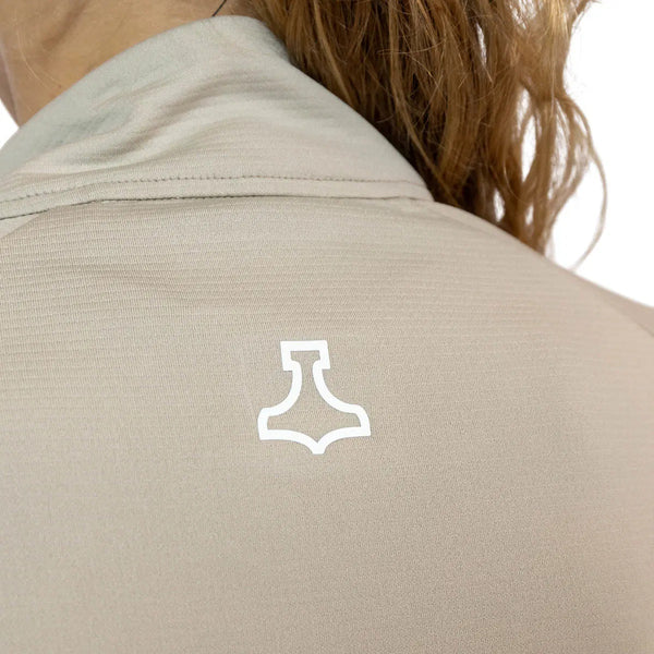 Nærbillede af en person iført en beige Liiteguard Ground-Tech 1/4 Zip, Dame, med et hvidt geometrisk logo under kraven. Det svedtransporterende stof ses, mens lysebrunt bølget hår falder blødt ned over ryggen.