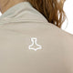 Nærbillede af en person iført en beige Liiteguard Ground-Tech 1/4 Zip, Dame, med et hvidt geometrisk logo under kraven. Det svedtransporterende stof ses, mens lysebrunt bølget hår falder blødt ned over ryggen.