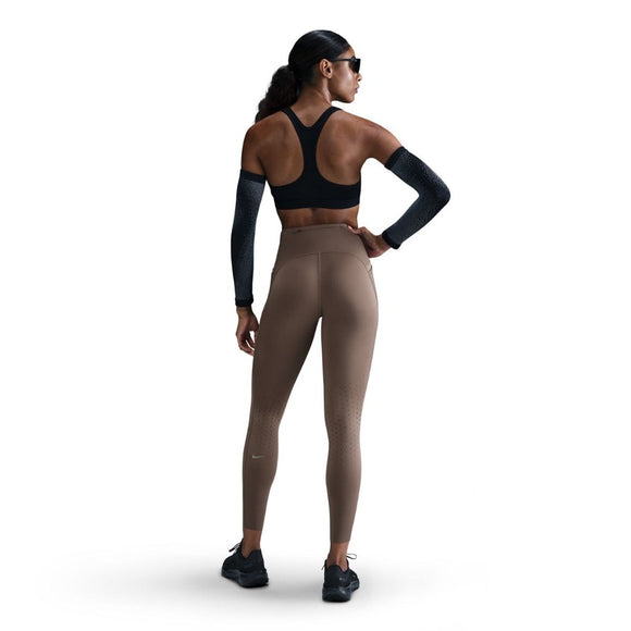En kvinde iført solbriller, sort sports-bh og Nike Swift High-Waisted 7/8 Running Tights med lommer i Mink Brown/Reflective Silver står med ansigtet vendt mod en almindelig hvid baggrund.
