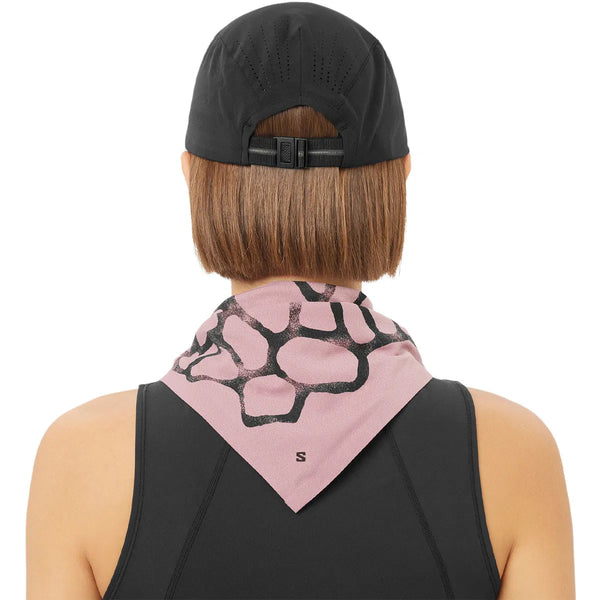 En person med kort brunt hår bærer en Salomon Shakeout Cap (Unisex, Deep Black) baglæns og en sort ærmeløs top med en pink bandana med et sort abstrakt mønster bundet om halsen. Billedet er taget bagfra.