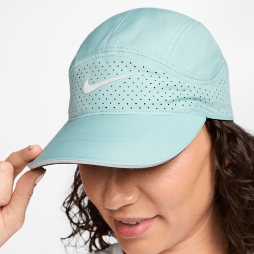 En person justerer kanten på en Nike Adv Fly Cap (Unisex, Cannon/Anthracite/Reflective Silv) med perforerede ventilationsåbninger og et hvidt Nike-logo; kun en del af ansigtet og hånden er synlige.