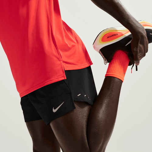 En person i orange skjorte, orange sokker og Nike Stride 5" Brief-Lined Running Shorts til mænd (Black/Reflective Silv) strækker sig ved at holde den ene fod tilbage. Kun underkroppen og benene vises; det fugttransporterende stof holder dem kølige.