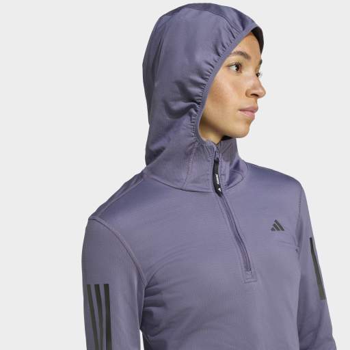 En kvinde iført Adidas Otr Bin Hz, Dame, Previo træningstrøje med AEROREADY-stof og en kvart lynlås foran samt sorte Adidas-logoer på bryst og ærme ser rigtig ud på en ensfarvet baggrund.