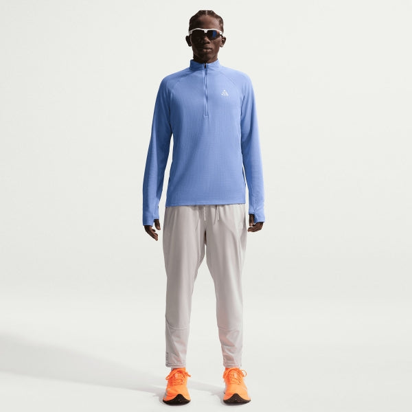 En mand står med front mod en almindelig hvid baggrund iført Nikes ACG Half-Zip Running Top til mænd i Royal Pulse/Summit White - kombineret med lysegrå bukser, knaldorange løbesko og solbriller for et ideelt løbetræningslook.