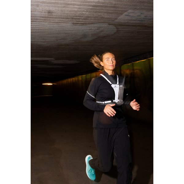 En person i sort tøj og blågrønne løbesko jogger gennem en mørk tunnel med tilbagestrøget hår og BeeSafe LED Harness USB Phone Pocket (Unisex, Silver) for at sikre løbeture i mørket.