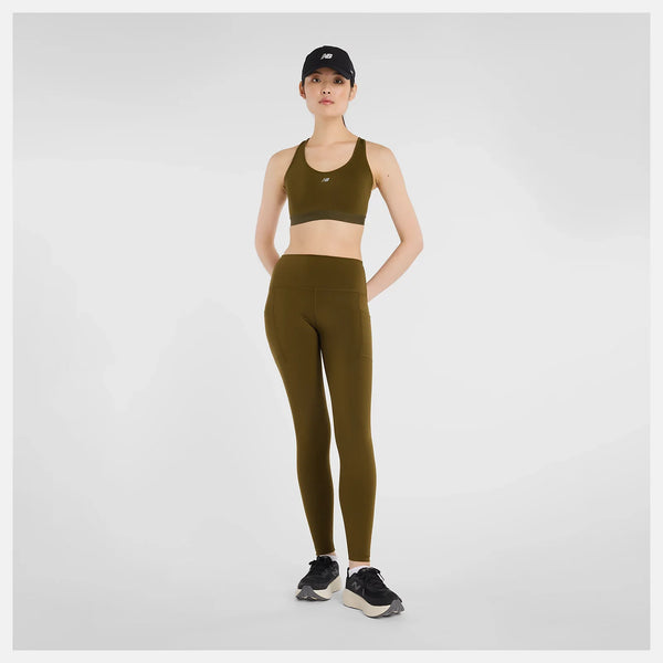 En kvinde ser fremad i New Balance's Sleek Pocket High Rise Legging i Dame Woodland, sammen med en matchende sports-bh, sorte sneakers og en sort kasket på en almindelig hvid baggrund.