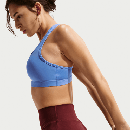 En kvinde iført Nike Swoosh Medium Sports Bra, Dame, Sapphire/White, og rødbrune leggings strækker armene bagud i profil mod en lys baggrund. Det svedtransporterende materiale holder hende komfortabel under træningen.