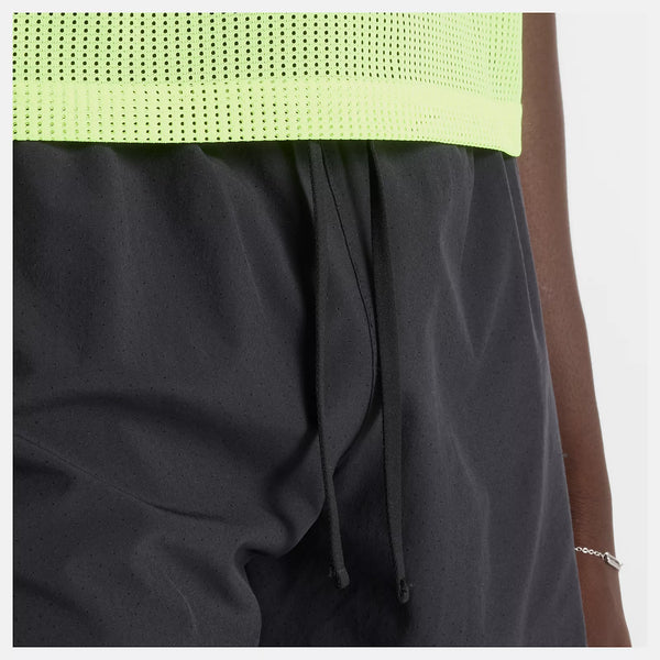 Et nærbillede viser en person iført sort New Balance RC Short 5", Herre, og en neongul perforeret mesh-top, hvor man kan se en del af armen og et tyndt armbånd.