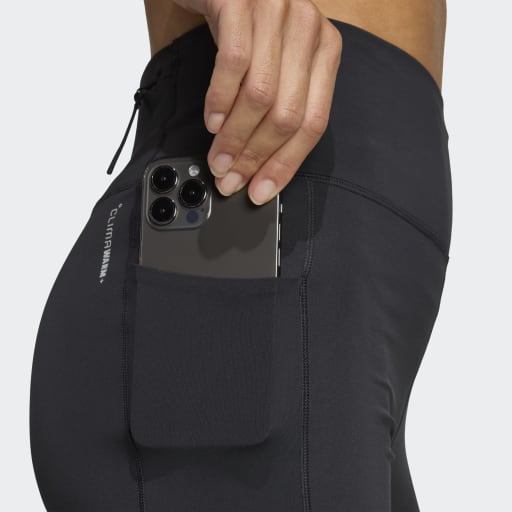 En person i sorte Adi365 Leggins, Dame by Adidas, stikker en smartphone med tre kameralinser ned i sidelommen med Adidas-logoet synligt nær lommen.