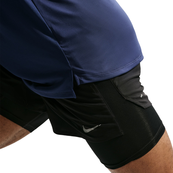 En person, der har en Nike Dri-FIT ADV langærmet løbetrøje i farvekombinationen Midnight Navy/Reflective Silver og sorte Nike-shorts på, er i færd med at strække ud. Billedet fremhæver den nedre del af overkroppen og lårene, med fokus på det markante Nike-logo på shortsene, set på en ensfarvet hvid baggrund.