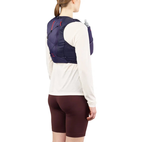 En person kigger væk, iført Salomon Active Skin 4 Set løbevest i Maritime Blue/Astral Aura/Haute Red over en lys langærmet skjorte og brune shorts. En vandflaske ligger i vestens lomme, klar til trailløbseventyr.