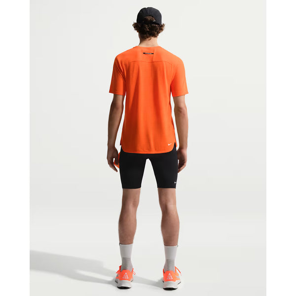 En person kigger væk fra kameraet iført Nike ACG Dfadv Solar Chase Ss herre i Safety Orange/Summit White, sorte shorts, grå sokker, orange løbesko og en sort kasket - ideel til udendørs træning på en almindelig lys baggrund.
