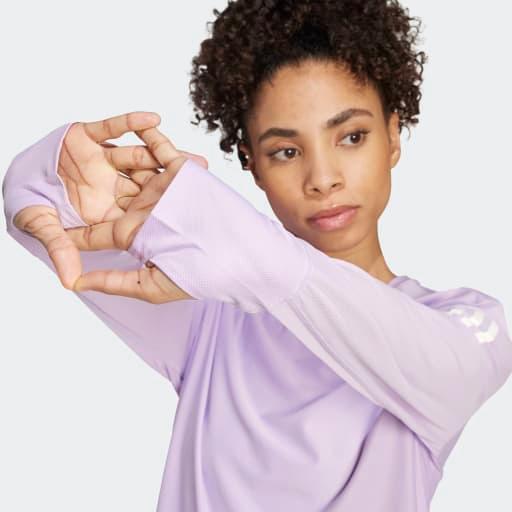 Iført Adidas Otr B Ls løbetrøje dame i powplu strækker en kvinde i en lyslilla langærmet træningstrøje med tommelfingerhuller sin venstre arm over brystet og kigger til siden mod en ensfarvet baggrund.