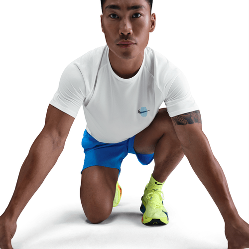 En mand i en Nike Stride Running Short-Sleeve Top (Herre, Sail), blå shorts og lysegule løbesko knæler på et knæ, klar til at sprinte, på en hvid baggrund.