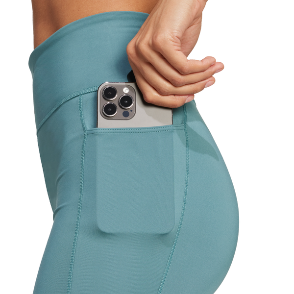 En kvinde iført Adidas OTR Short Tights 6" i PRETEA putter en smartphone med tre kameralinser i sidelommen på de blågrønne leggings.