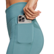 En kvinde iført Adidas OTR Short Tights 6" i PRETEA putter en smartphone med tre kameralinser i sidelommen på de blågrønne leggings.