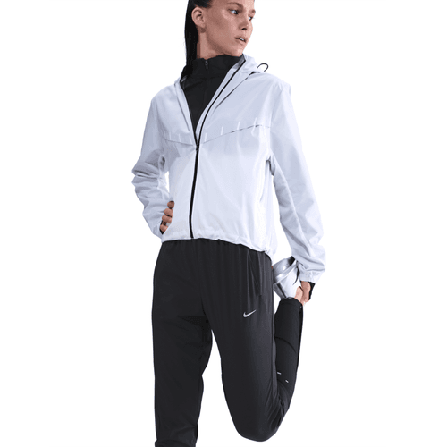 En person i hvid jakke med lynlås og sorte Nike Dri-FIT Mid-Rise Running Pants (Dame, Black/Reflective Silv) strækker benet, og den hvide baggrund fremhæver komforten og stilen i dette træningstøj dame-look.