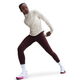 En kvinde poserer atletisk mod en hvid baggrund iført Nikes Therma-FIT Turtle-Neck Running Top i Sail/Reflective Silver, parret med rødbrune leggings, hvide sokker og hvide Nike-sneakers med pink såler.