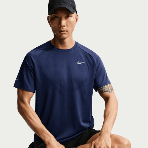 En person iført Nike Stride SS Running Top, Herre, Midnight Navy/Reflective Silv, sorte shorts og en sort kasket sidder med front mod en lys baggrund med et neutralt udtryk og synlige tatoveringer på armene.