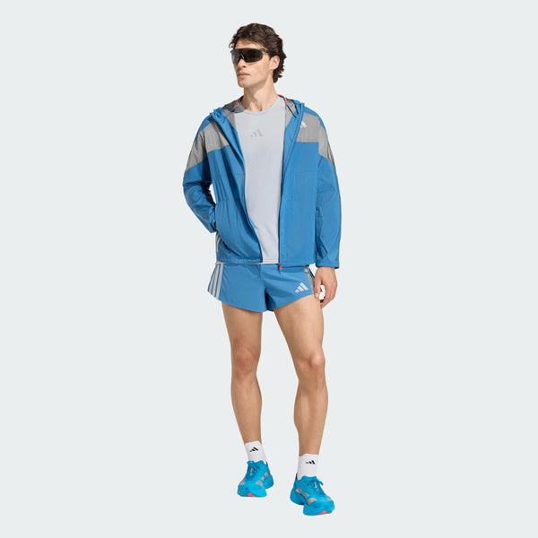 En person står på en lys baggrund iført Adidas Adizero Hakone Ekiden Shorts Herre Duinn, en blå og grå jakke, en lysegrå skjorte, blå løbesko, hvide sokker og mørke solbriller.