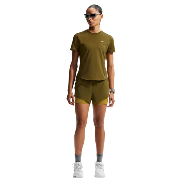 En kvinde kigger frem iført solbriller, en olivengrøn sportstop, Nike Nk Swift Df Mr 2N1 Shrt Dame shorts i Olive Flak/Peat Moss/Reflective Silv, grå sokker, hvide sneakers, et ur og har håret trukket tilbage.