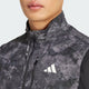 Nærbillede af en person iført Adidas Otr Sd Vest M til mænd i Grey Four/Black med lynlås og logo på brystet. Den svedtransporterende vest er ideel til aktive dage. Ansigt delvist synligt, ensfarvet baggrund.
