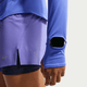En person i lilla løbeshorts og en Nike Swift UV Long Sleeve Shirt til kvinder i Sapphire/Reflective Silver med et urvindue på ærmet, der afslører et smartwatch. Baggrunden er ensfarvet og lys.