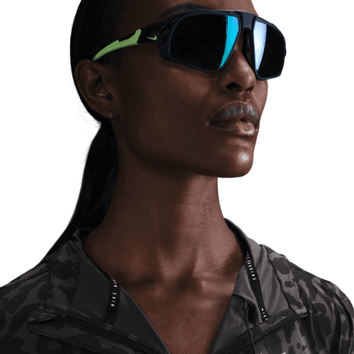 En person bærer overdimensionerede sorte sportssolbriller med grønne detaljer og en Nike Dri-FIT løbejakke til kvinder i sort/antrazit/reflekterende sølv, mens han kigger lidt opad mod en hvid baggrund.