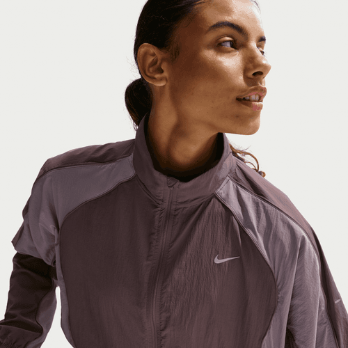 En person iført Nike Swift Repel UV Protection Running Jacket til kvinder i Tattoo/LT Violet Ore/Reflective Silver med lynlås i kraven kigger til siden med håret bundet tilbage mod en ensfarvet baggrund.
