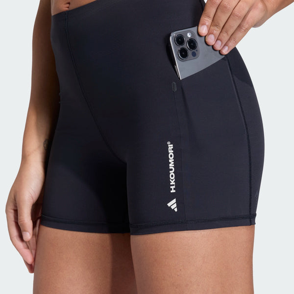 En kvinde er iført sorte Adi365 Hk Tight shorts fra Adidas med en telefon i sidelommen. "HKOUNORI" og et lille geometrisk logo er trykt på venstre ben, så det ligner træningstights kvinder.