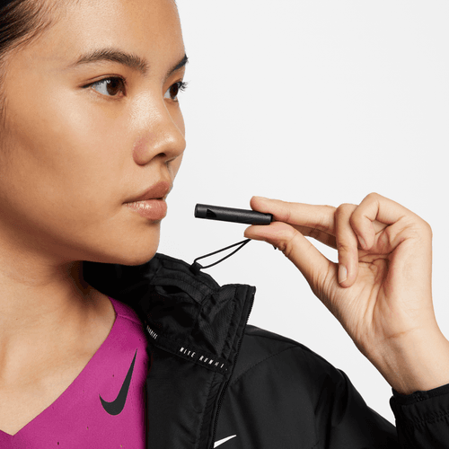 En person iført en sort Nike Therma-FIT løbejakke, Dame, Black/Reflective Silv og en lilla skjorte holder en sort fløjte ved munden og kigger til siden.