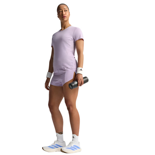 En kvinde holder en vandflaske i sin højre hånd, iført Adidas Adizero E Tee til kvinder i Powplu, sammen med hvide armbånd og hvide sneakers med lilla detaljer. Hun kigger opad med et neutralt udtryk.