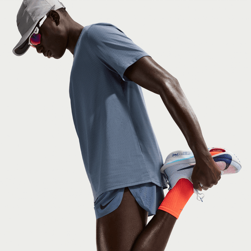 En person i sportstøj, herunder en grå kasket, blå skjorte og Nike AeroSwift 4In Brief-Lined Running Shorts for herrer i Diffused Blue/Black, strækker sit ben ved at holde foden bag sig mod en almindelig hvid baggrund.