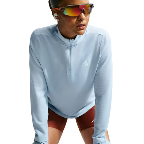 En person i Nike ACG Half Zip Running Top, Dame, Hydrogen Blue/Summit White, røde sportsshorts og reflekterende solbriller læner sig frem med begge hænder synlige mod en almindelig hvid baggrund.