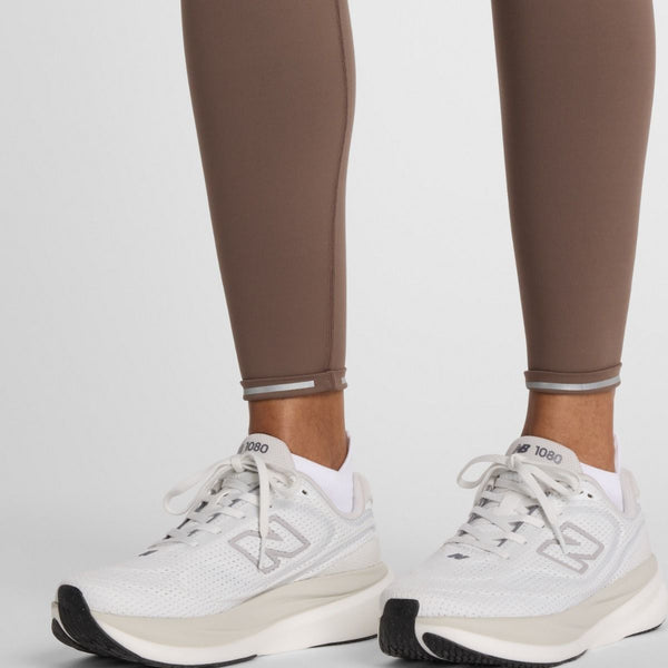 En person er iført Sleek Pocket High Rise Legging 27, Dame, Cortado og hvide New Balance-atletiksko og står på en lys overflade, hvor kun underben og fødder er synlige.