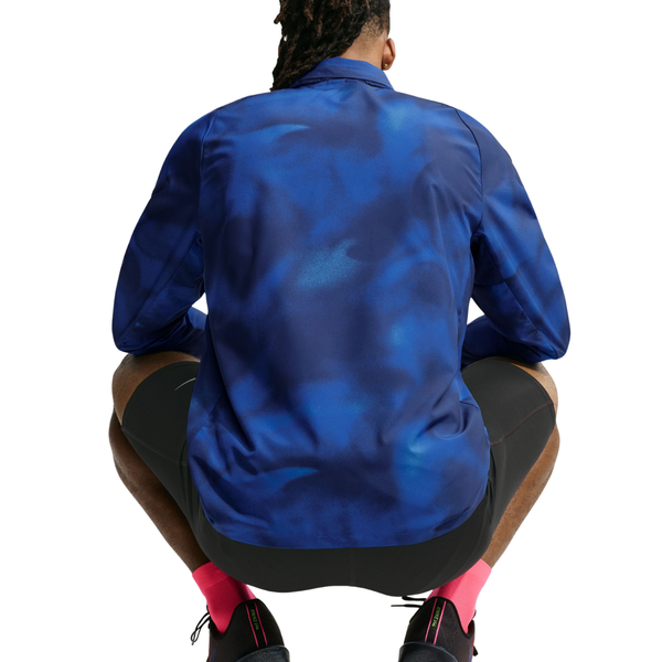 En person med flettet hår, iført Nike Miler UV Repel-løbejakken til mænd i farven Midnight Navy/Reflective Silver, sorte shorts og lyserøde sokker, sidder i hug med ryggen til kameraet foran en hvid baggrund.