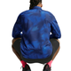 En person med flettet hår, iført Nike Miler UV Repel-løbejakken til mænd i farven Midnight Navy/Reflective Silver, sorte shorts og lyserøde sokker, sidder i hug med ryggen til kameraet foran en hvid baggrund.