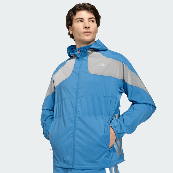 En mand i en blå og grå Adidas Adizero Hakone Ekiden Jacket, Herre, Duinn/Sliver - en ultralet løbejakke med lynlåslommer - står med hænderne i lommerne og kigger til siden mod en almindelig lys baggrund.