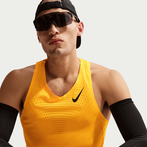 En person i en gul Nike AeroSwift Running Singlet, Herre, Laser Orange/Black, med sorte ærmer, store sorte solbriller og en bagudvendt sort kasket ser fremad mod en almindelig lys baggrund.