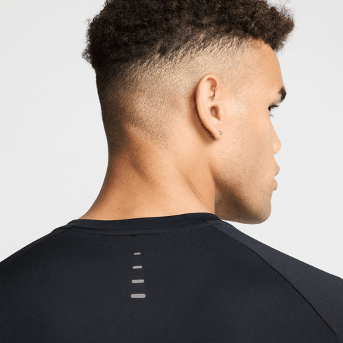 En mand med kort, krøllet hår og fade ses bagfra og fra siden iført Nike Stride SS Running Top (Herre) i Black/Reflective Silv med fire små vandrette refleksstriber på den øverste del af ryggen.